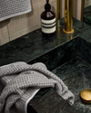 Grey fog hand towel draped casually on a dark green marble countertop with brass fixtures and hand soap.