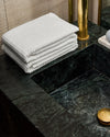 White hand towels stacked on a dark green marble countertop with brass fixtures and a gold dish.