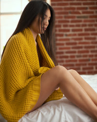 A woman wrapped in the ONSEN Ochre Waffle Bath Sheet in a bedroom setting.