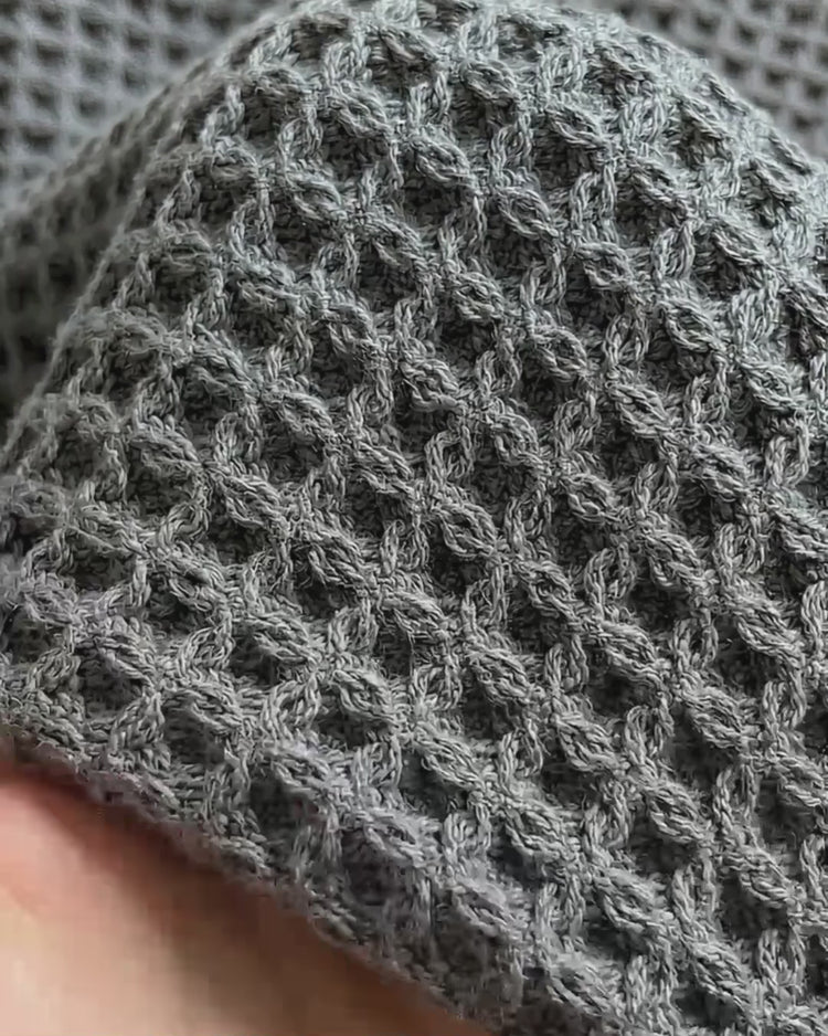 A close-up macro shot of a textured cinder grey waffle-weave bath towel being held.