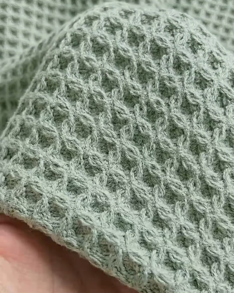 A close-up macro shot of a textured sage green ONSEN waffle-weave bath towel being held.