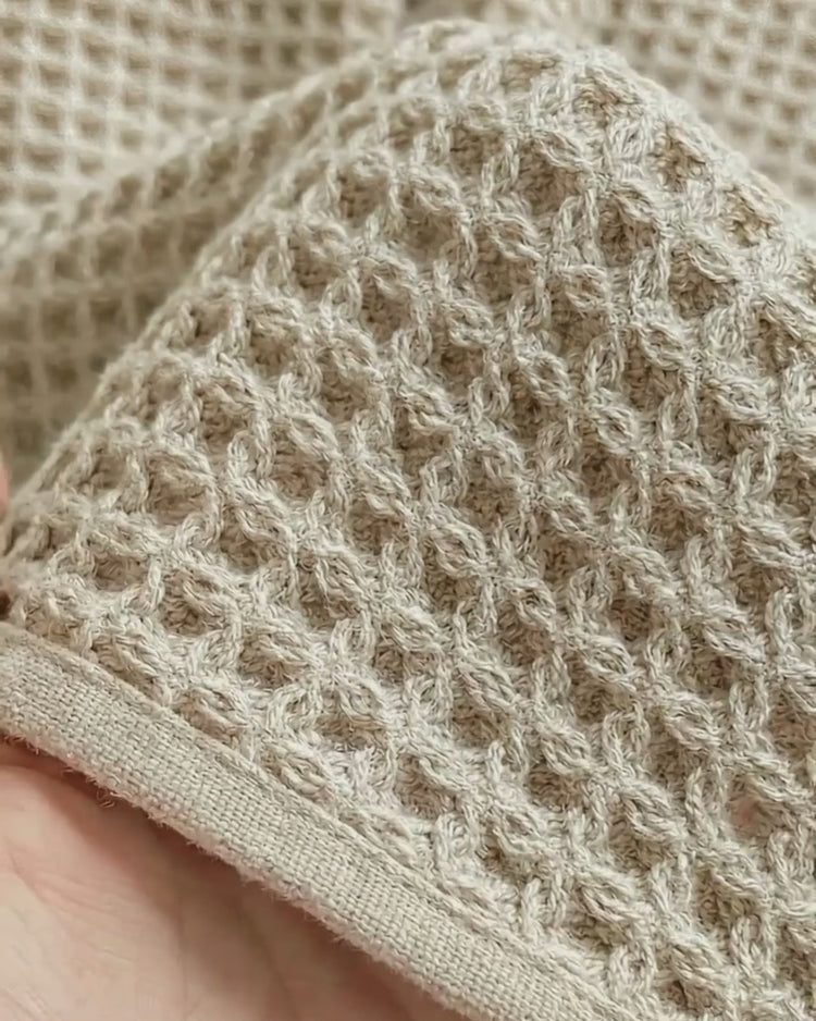 A close-up of a textured beige or oatmeal-colored waffle-weave bath towel showing its intricate fabric pattern and a smooth stitched border.